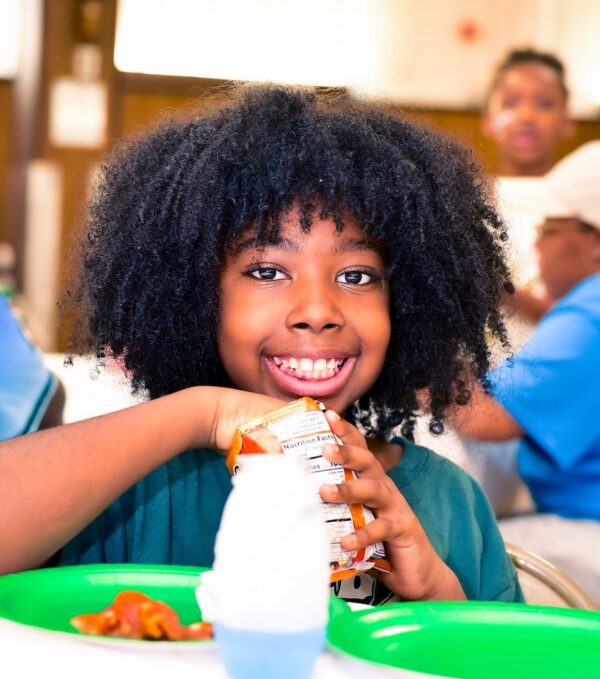 A girl is sitting in the Genesis School cafeteria, eating chips, smiling at the camera
